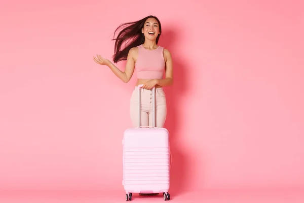 Ταξίδια, διακοπές και διακοπές έννοια. Full-length of carefree sassy brunette Asian girl επιτέλους πάει στο εξωτερικό, περπατώντας κατά μήκος του αεροδρομίου, πετώντας τα μαλλιά και μεταφέρουν βαλίτσα πάνω από ροζ φόντο — Φωτογραφία Αρχείου