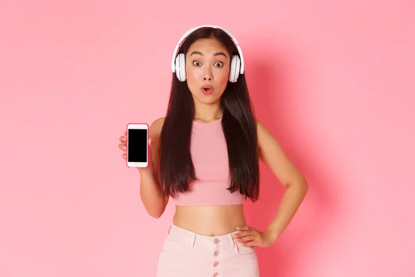 Tecnologia, comunicação e conceito de estilo de vida online. Impressionado menina asiática encontrado podcast incrível, mostrando tela do smartphone como usar fones de ouvido, ouvir música, fundo rosa — Fotografia de Stock