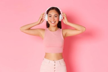 Güzel görünümlü Asyalı bir kızın portresi kulaklık taktı ve gülümsedi. Kulaklık takan kadın ilginç bir podcast dinlemeye başladı. Pembe arka plan, canlı uygulamadan müziğin keyfini çıkaran bir kadın.