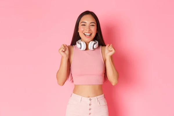 Retrato de alegre chica asiática optimista en ropa de moda, con auriculares alrededor del cuello, saltando de la felicidad o la anticipación, regocijándose por las grandes noticias, ganando algo, fondo rosa — Foto de Stock