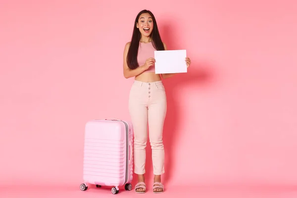 Reisen, Urlaub und Urlaubskonzept. Volle Länge von aufgeregten und überraschten, ziemlich asiatischen Touristin, Mädchen mit einem Stück Papier mit Ihrem Zeichen, steht in der Nähe Koffer über rosa Hintergrund — Stockfoto