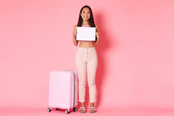 Reisen, Urlaub und Urlaubskonzept. Ganzer Länge schöne lächelnde asiatische Touristin mit Koffer über rosa Hintergrund, wartet auf jemanden Flughafen, mit Zeichen auf weißem Papier — Stockfoto