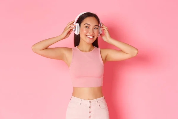 Πορτρέτο της κομψής όμορφη ασιατική κορίτσι τεθεί σε ακουστικά και χαμογελώντας. Η γυναίκα στα ακουστικά αρχίζει να ακούει ενδιαφέρον podcast. Γυναίκα απολαμβάνοντας μουσική από streaming app, ροζ φόντο — Φωτογραφία Αρχείου
