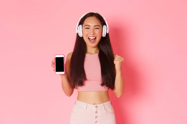 テクノロジー、コミュニケーション、オンラインライフスタイルの概念。華やかなダンスアジアの女の子は、アップビートを見て、ヘッドフォンで音楽を聞いて、スマートフォンの画面、ピンクの背景を示す喜び — ストック写真