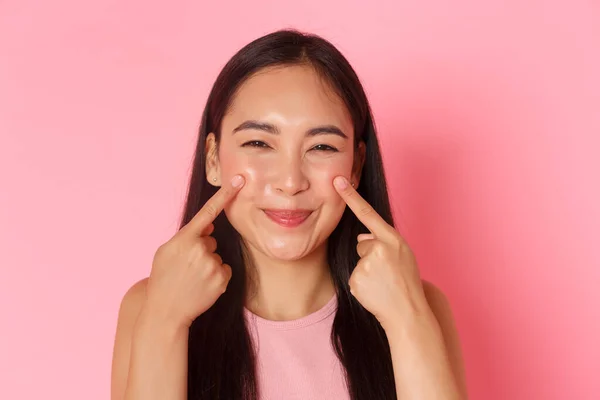 Beauty, fashion and lifestyle concept. Close-up of silly and cute, adorable asian girl poking her cheeks and smiling happily, promo of skincare or makeup product, pink background