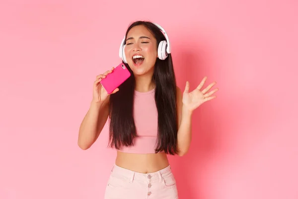 Pas-up portret atrakcyjnej stylowej azjatyckiej dziewczyny zamknij oczy i śpiewając wzdłuż piosenki podczas słuchania muzyki w słuchawkach, grając w grę karaoke na telefon komórkowy, stojąc nad różowym tle — Zdjęcie stockowe