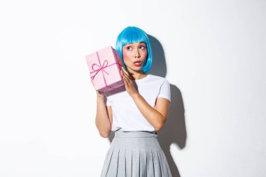 Mavi peruklu şirin bir Asyalı kız hediye kutusunu sallıyor, içinde ne olduğunu merak ediyor, beyaz arka planda duruyor.