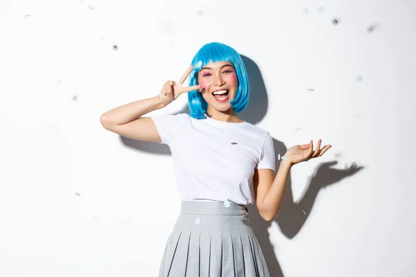 Retrato de chica asiática feliz en peluca azul celebrando halloween, lanzando confeti y mostrando gesto de paz kawaii, de pie sobre fondo blanco — Foto de Stock