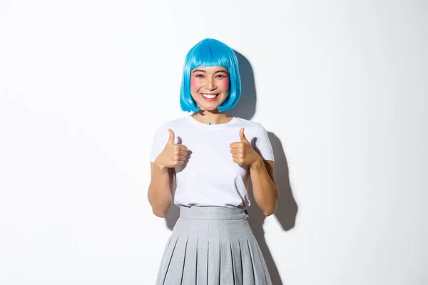 Retrato de menina sorridente satisfeito em peruca azul e traje de Halloween, mostrando polegar-se em aprovação, como algo bom, de pé sobre fundo branco — Fotografia de Stock