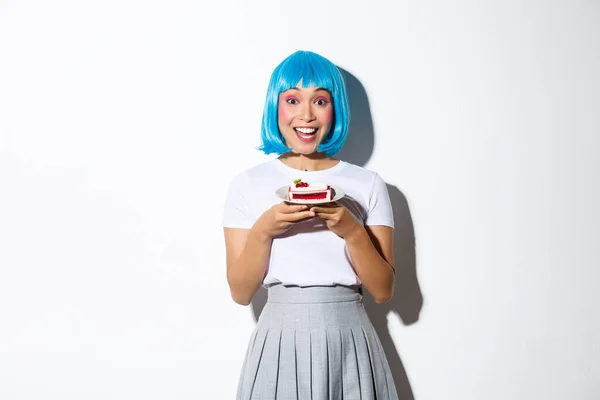 Ritratto di felice sorridente ragazza asiatica con trucco carino, indossa parrucca corta blu, tenendo pezzo di torta, in piedi su sfondo bianco — Foto Stock