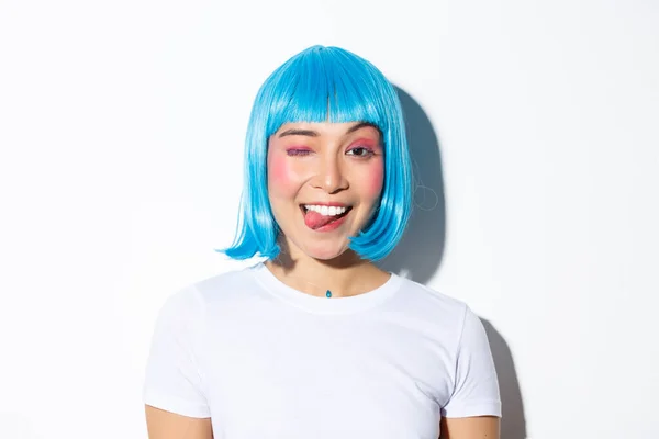 Крупный план привлекательной тусовщицы в голубом парике, показывающей язык и подмигивающей счастливой, празднующей Хэллоуин, стоящей на белом фоне — стоковое фото