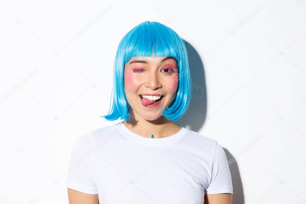 Close-up of attractive party girl in blue wig, showing tongue and winking happy, celebrating halloween, standing over white background
