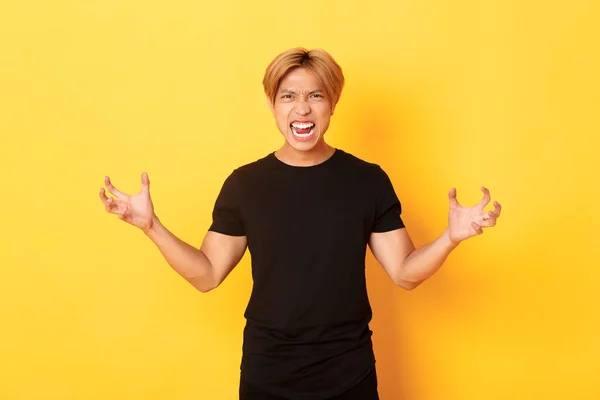 Portrait of outraged, pissed-off asian man shaking hands hateful and grimacing, shouting angry, standing over yellow background — стоковое фото