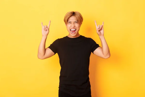 Portré boldog jóképű ázsiai férfi szórakozás és bulizás, bemutató rock-n-roll gesztus, álló sárga háttér — Stock Fotó