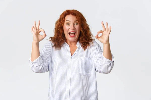 Excited and happy, lucky middle-aged redhead woman reacting to amazing news, showing okay gesture and smiling upbeat, recommend excellent tour, great prices in shop, white background — Stock Photo, Image