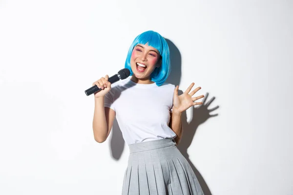 Cheerful cute asian girl dressed up as anime character for halloween party, wearing blue wig and schoolgirl costume, holding microphone and singing karaoke, standing over white background