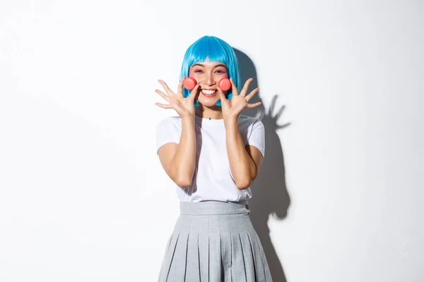 Obraz pięknej uśmiechniętej azjatyckiej dziewczyny wyglądającej głupio, pokazującej makaroniki, noszącej niebieską perukę anime, stojącej na białym tle — Zdjęcie stockowe