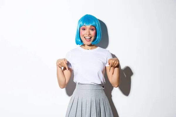 Obraz szczęśliwej głupiej azjatyckiej dziewczyny pokazującej logo lub reklamę produktu, pokazującej palce w dół i uśmiechniętej, noszącej niebieską krótką perukę, stojącej nad białym tłem — Zdjęcie stockowe