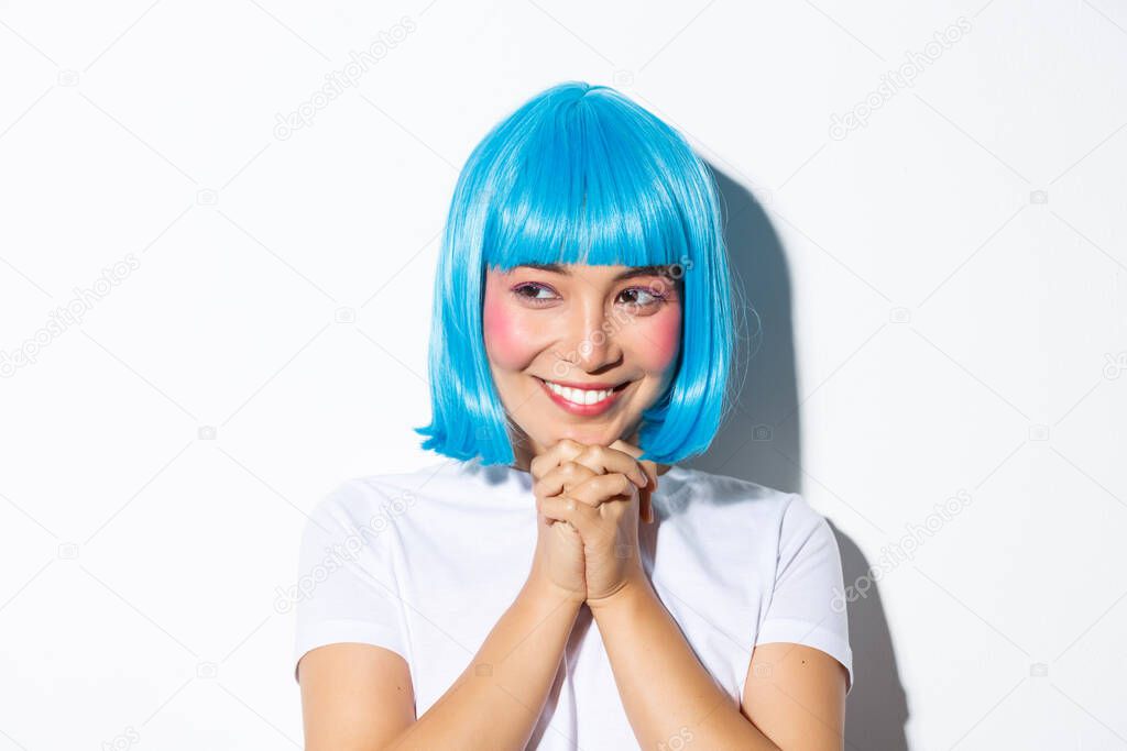 Close-up of adorable dreamy asian girl looking left and smiling, clasping hands together hopeful, wearing blue wig for halloween party, standing over white background