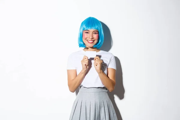 Retrato de linda chica asiática en peluca azul mostrando tarjeta de crédito, sonriendo feliz, de pie sobre fondo blanco — Foto de Stock
