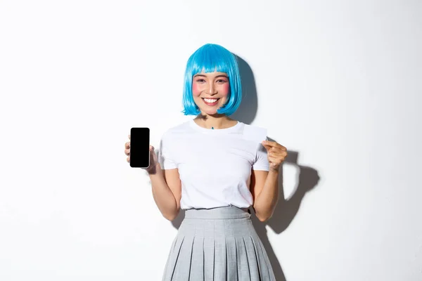 Image of cute asian girl dressed up as anime character in blue wig, holding credit card and showing mobile phone screen, standing over white background