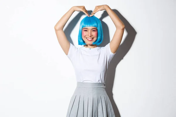 Porträt eines niedlichen japanischen Mädchens mit blauer Perücke, das eine Herzgeste zeigt und über weißem Hintergrund lächelt — Stockfoto