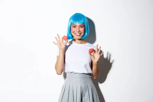Retrato de kawaii chica asiática con maquillaje glamour, vistiendo peluca azul, mostrando macarrones y sonriendo, de pie sobre fondo blanco — Foto de Stock