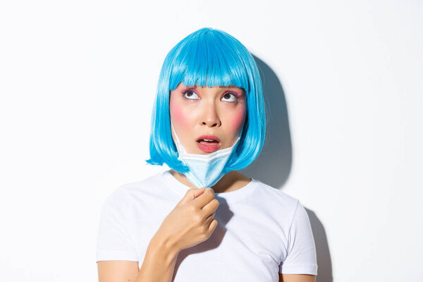 Concept of halloween celebration and coronavirus. Close-up of confused asian woman in blue wig, taking-off medical mask and looking up, standing over white background