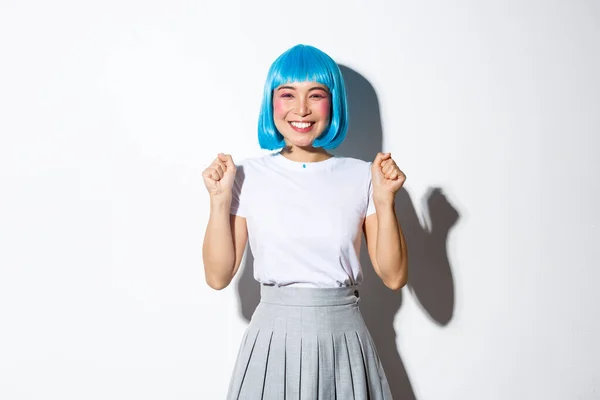 Retrato de la alegre chica asiática kawaii en peluca de fiesta azul celebrando la victoria, sonriendo feliz y saltando de alegría, triunfando sobre el éxito, de pie sobre fondo blanco — Foto de Stock
