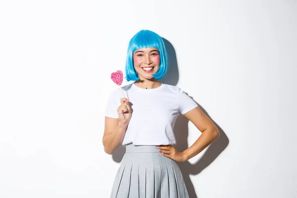 Imagen de linda chica asiática en peluca azul sonriendo feliz, sosteniendo dulces, yendo truco o trato en Halloween en su traje de colegiala, de pie sobre fondo blanco — Foto de Stock