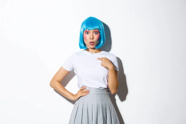 Obraz zaskoczonej azjatyckiej dziewczyny dyszącej zdumiony i wskazując palcem na siebie, nosząc niebieską perukę na halloween party, stojąc nad białym tle — Zdjęcie stockowe