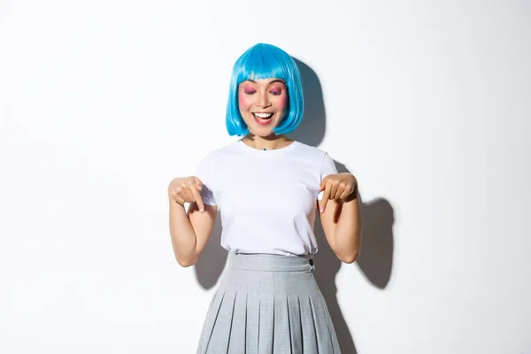 Entuzjastyczna piękna azjatycka dziewczyna w niebieskiej peruce, kostium halloween, wskazując palcami w dół z uśmiechem zadowolony, pokazując reklamy, białe tło — Zdjęcie stockowe