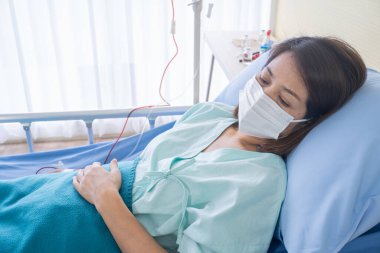 Asyalı bir kadın hasta, yüksek ateşi var, hapşırıyor, hastanın elbisesinin içinde iyileşiyor. Hastanedeki hasta yatağında yüz maskeleri virüsü koruyor. covid19 konsepti