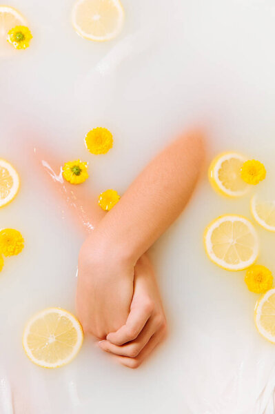 Woman's crossed hands in water and milk bathtub, with slices of lemon and yellow flowers. Relaxing, Fresh SPA and beauty concept. 