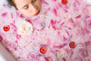 Kırmızı dudaklı güzel kız küvette yatıyor su, süt, şakayık ve mumlarla