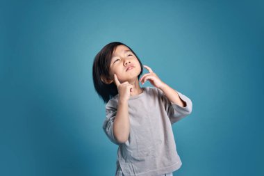 Güzel Asyalı küçük çocuk merak gösterir, hayal eder, düşünür ya da tereddüt eder. Stüdyodaki boş alan, renkli mavi arka planda izole edilmiş. Okul için eğitim kavramı.