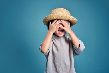 Mutlu Asyalı çocuk iki eliyle yüzünü kapatıyor. Stüdyodaki boş alan, renkli mavi arka planda izole edilmiş. Okul için eğitim kavramı.