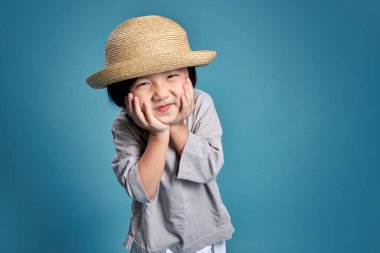Mutlu Asyalı çocuk şapkasıyla gülümseyip iki eliyle yüzünü tutuyor. Stüdyodaki boş alan, renkli mavi arka planda izole edilmiş. Okul için eğitim kavramı.