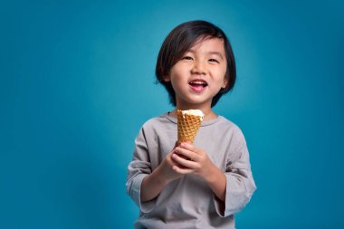 Mutlu gülümseyen Asyalı küçük çocuk elinde lezzetli bir dondurma tutuyor. Stüdyodaki boş alan, renkli mavi arka planda izole edilmiş. Okul için eğitim kavramı.