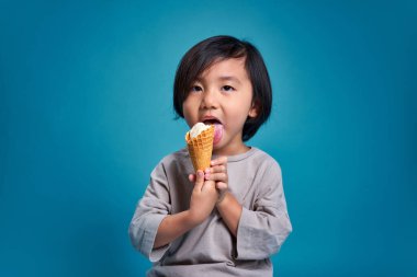 Mutlu Asyalı küçük çocuk lezzetli dondurmaları yalıyor. Stüdyodaki boş alan, renkli mavi arka planda izole edilmiş. Okul için eğitim kavramı.