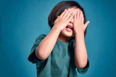 Mutlu Asyalı küçük çocuk iki eliyle yüzünü kapatıyor. Stüdyoda boş bir alan var. Renkli mavi arka planda izole edilmiş. Okul için eğitim kavramı.