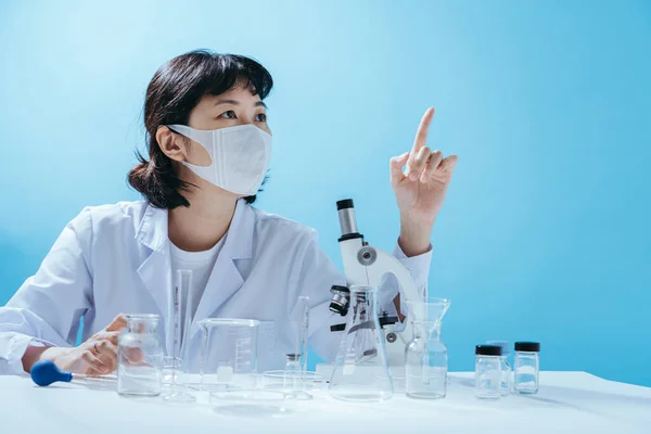 Smart young asian female scientist in lab coat with Medical face mask pointing. Empty space in studio shot isolated on colorful blue background. Medical concept.