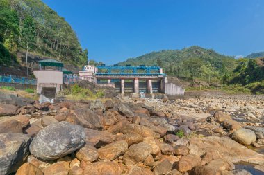 DOOARS, Batı BENGAL, INDIA - 6 ARALIK 2017 Joldhaka nehir projesi - Joldhaka nehri kıyısındaki hidroelektrik santrali. Önplanda nehir kıyısının taşları ve arkada mavi gökyüzü.