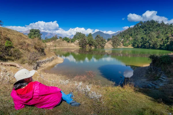 Yalnız gezgin, bekar bayan turist, Deoria Tal 'in güzelliğinin tadını çıkarırken yeşil çimlerin üzerinde, Uttarakhand, Hindistan' da yüksek irtifada uzanan göl. Arkaplanda karla kaplı dağlar olan mavi gökyüzü.