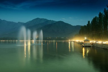 Dal Gölü üzerindeki fıskiyeler, arka planda dağlar, alacakaranlıkta. Dal Gölü, Hindistan 'ın Jammu ve Kashmir' in en ünlü gölleridir ve dünyanın dört bir yanından gelen bir turistik merkezdir..