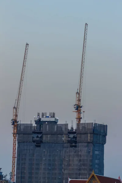 Luffing jib tower crane at condominium construction site over steel framework among high rise building. Skyscraper building construction with the tower cranes on top under the dramatic sky background.