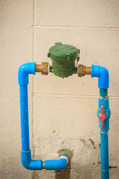 Vintage water meter installed with bronze joint, PVC elbow connector, blue PVC pipe and red water valve on the grungy wall.