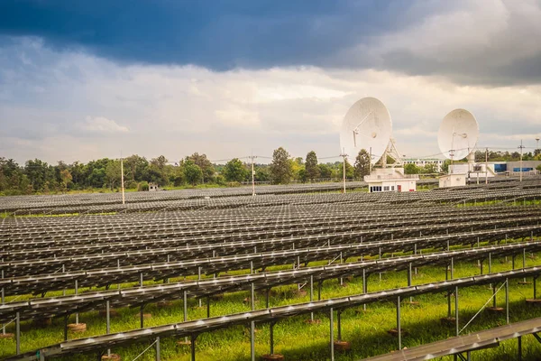 Large scale solar farm with the satellite dishes under dramatic blue and cloudy sky background. Mega photovoltaic power plant in green grass field with two satellite dishes under stormy sky background