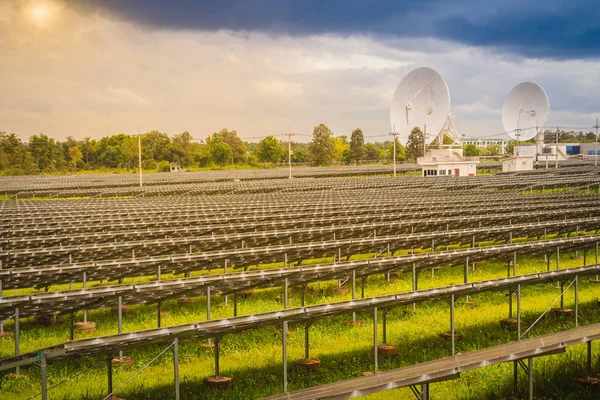 Large scale solar farm with the satellite dishes under dramatic blue and cloudy sky background. Mega photovoltaic power plant in green grass field with two satellite dishes under stormy sky background