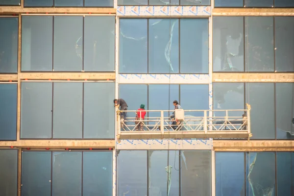 Construction workers on a suspended cradle platform on a skyscraper glass facade. Suspended cradle is similar to temporary suspended scaffolds for workers to work outside the skyscraper building.